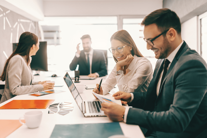 Dos colegas alegres con ropa formal resolviendo problemas y mirando papeleo. En segundo plano, empleados hablando. Es literalmente cierto que se logra mejor y más rápido ayudando a otros a tener éxito.