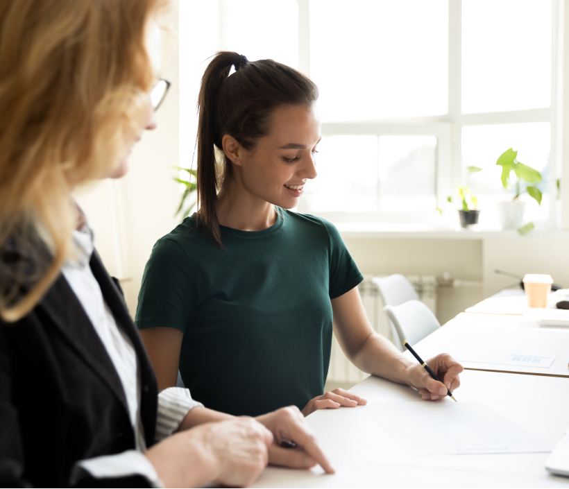 dos chicas de perfil, una morena con una coleta con una camiseta verde y está escribiendo, y una mujer rubias está señalando el papel con un dedo, ambas visten elegantes y parece que están en una oficina de consultoría